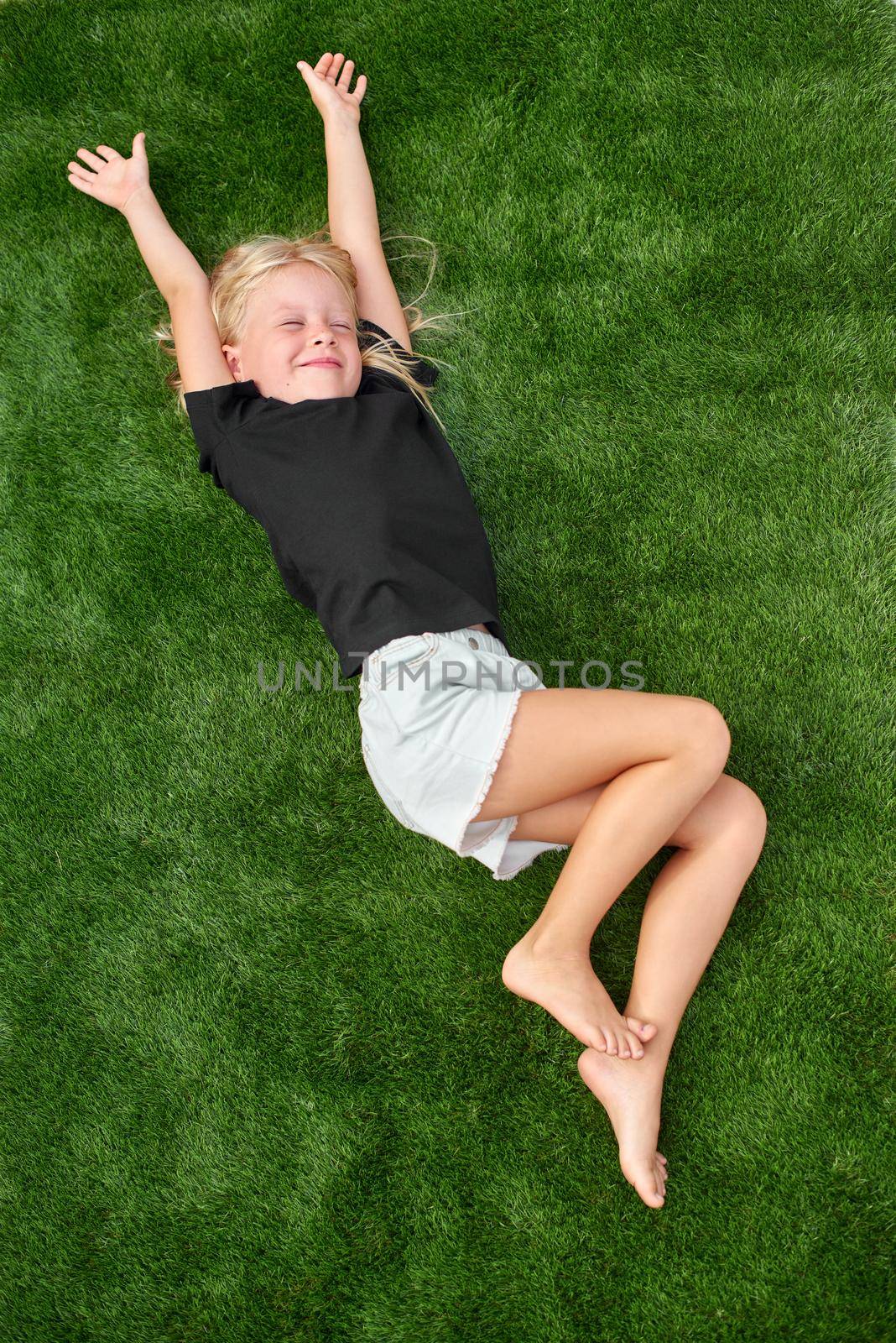 Top view. Mock up. Cute blonde child girl with closed eyes lying, relaxing, stretching on green grass. Smiling preschool girl 5 - 6 years old in black t shirt. Summer vacation. Leisure. People concept