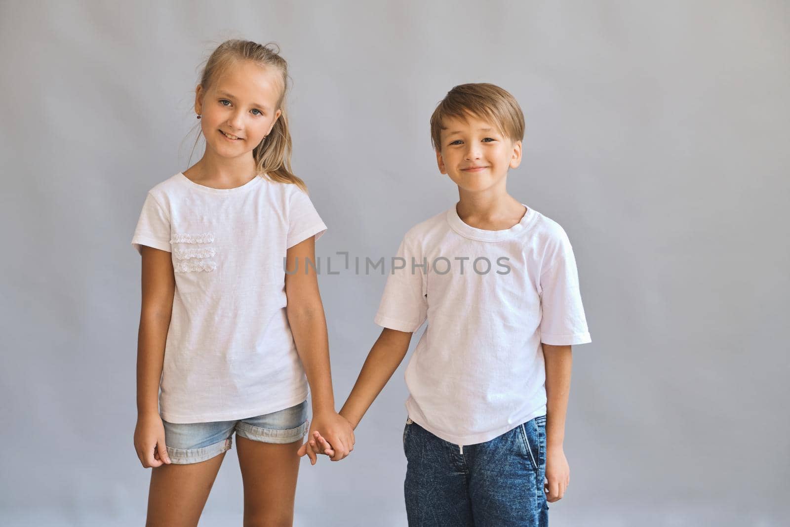 Cute smilling two kids, little boy and girl in white t-shirts hold hands best friends on gray background. Blank white t-shirt for your design