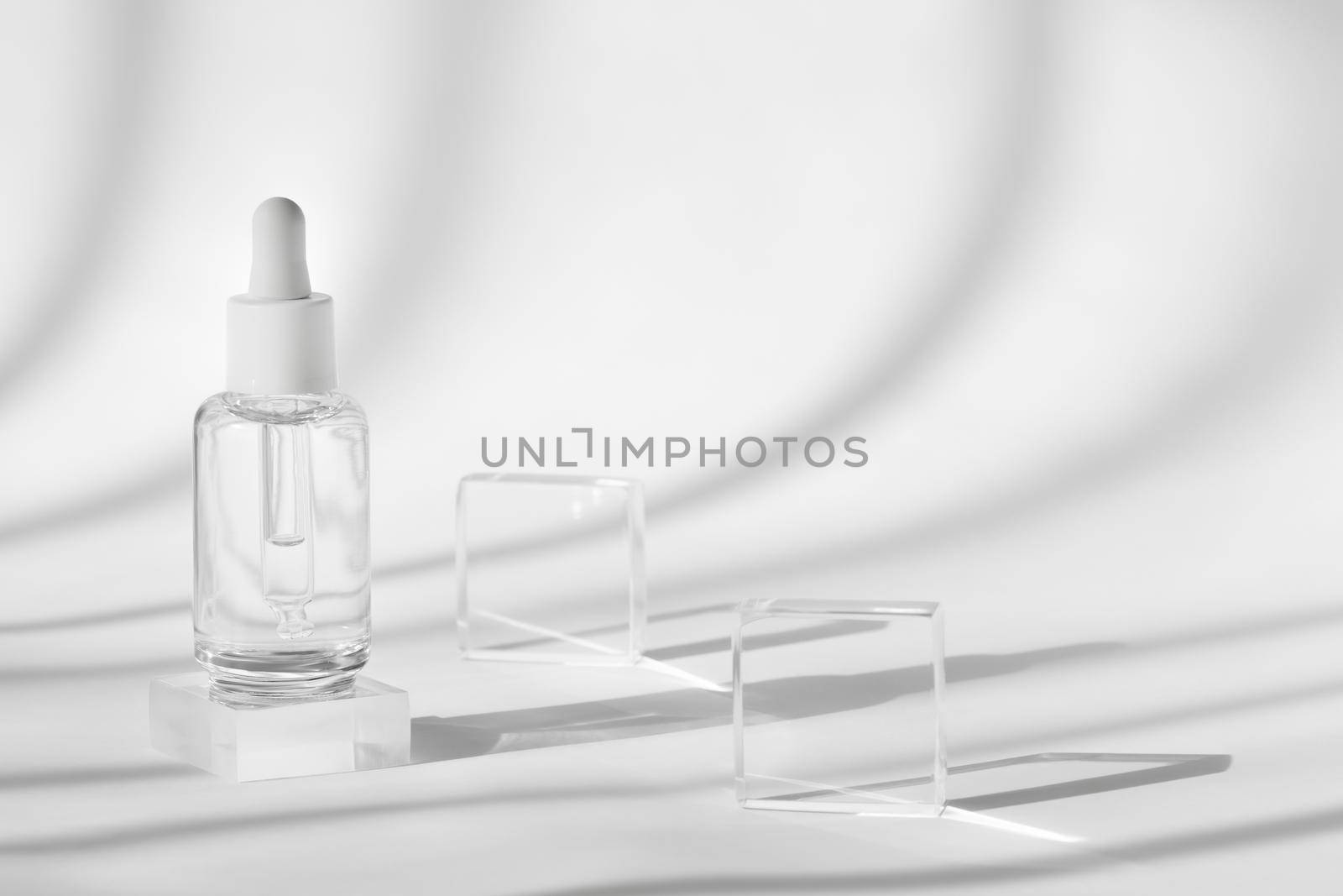 Serum cosmetic bottle mockup on acrylic transparent solid block pedestal on white background, shadow from sun, natural light from windows. Geometric stand, podium for cosmetics, product presentation