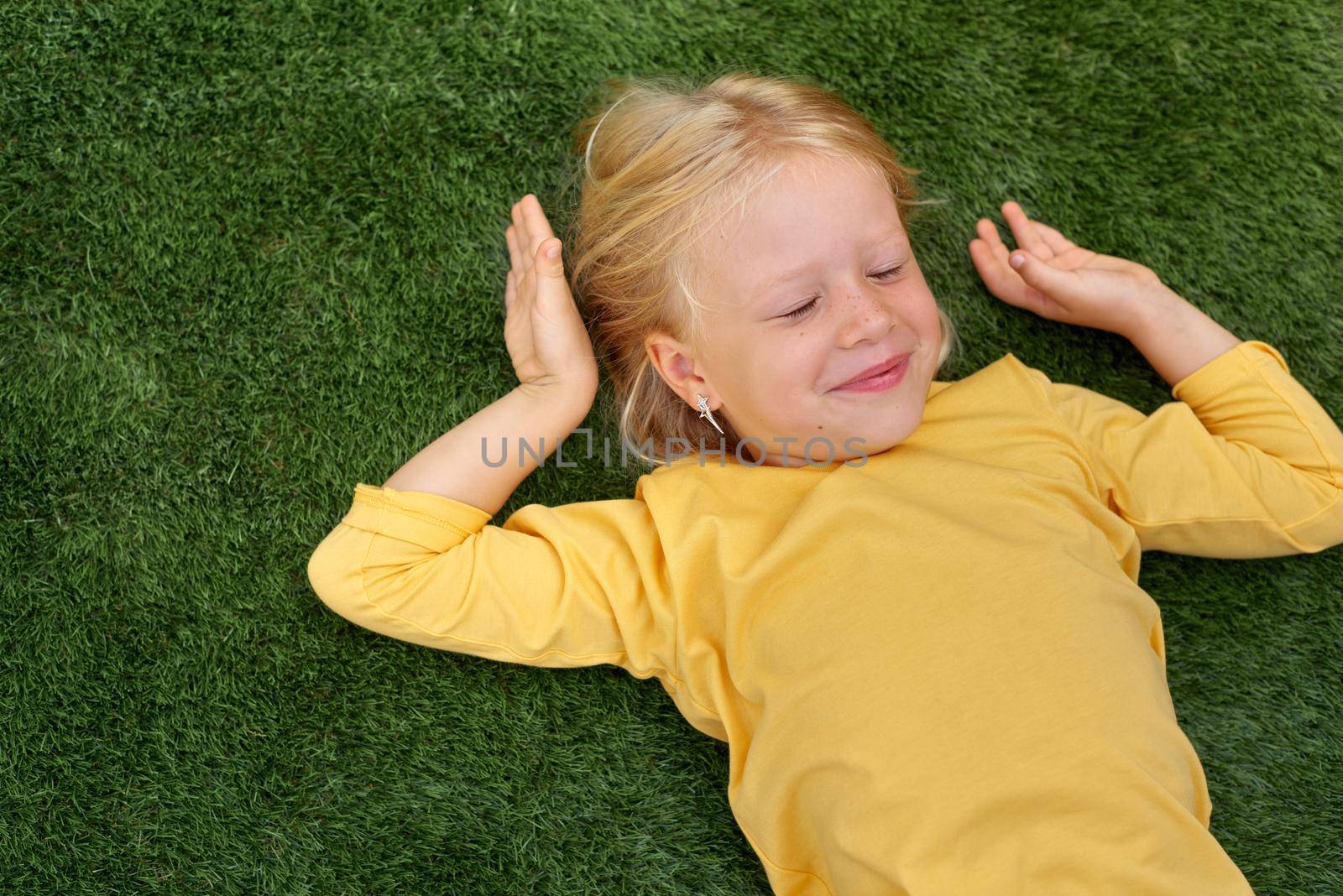 Top view. Mock up for logo, text, design. Blonde long hair child girl lying stretching on green grass. Preschool girl 5-6 years old in yellow t shirt. Lifestyle Summer vacation Leisure. People