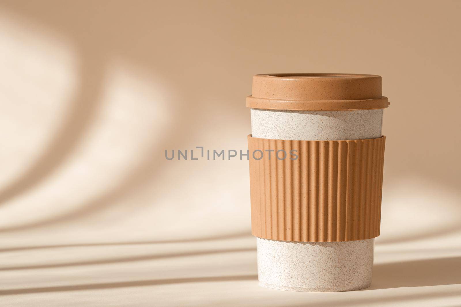 Reusable cup, biodegradable travel plastic coffee mug for take away. Sustainable bamboo eco friendly cup with silicone holder on natural shadow beige background. Zero waste, sustainability concept.