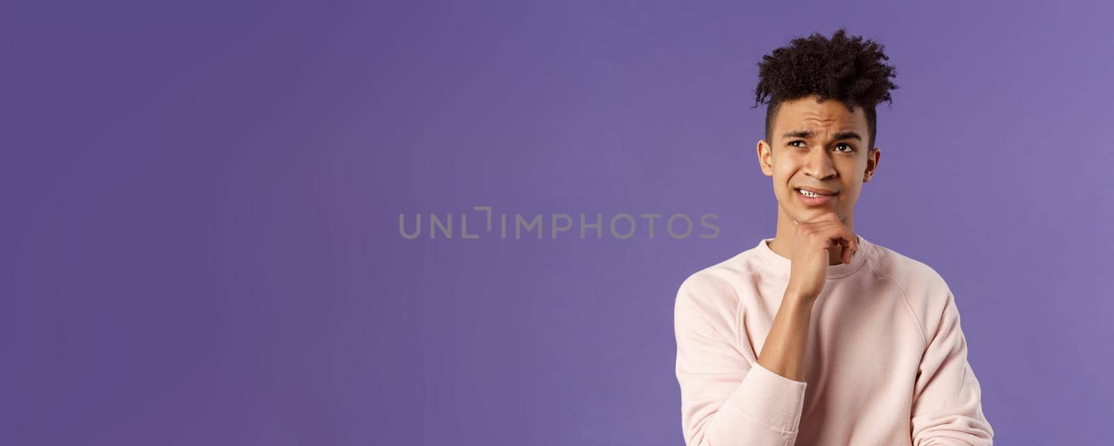 Close-up portrait of indecisive, puzzled young man facing difficult choice, look up thoughtful, touch chin and grimacing while thinking, making-up idea or decision, purple background.