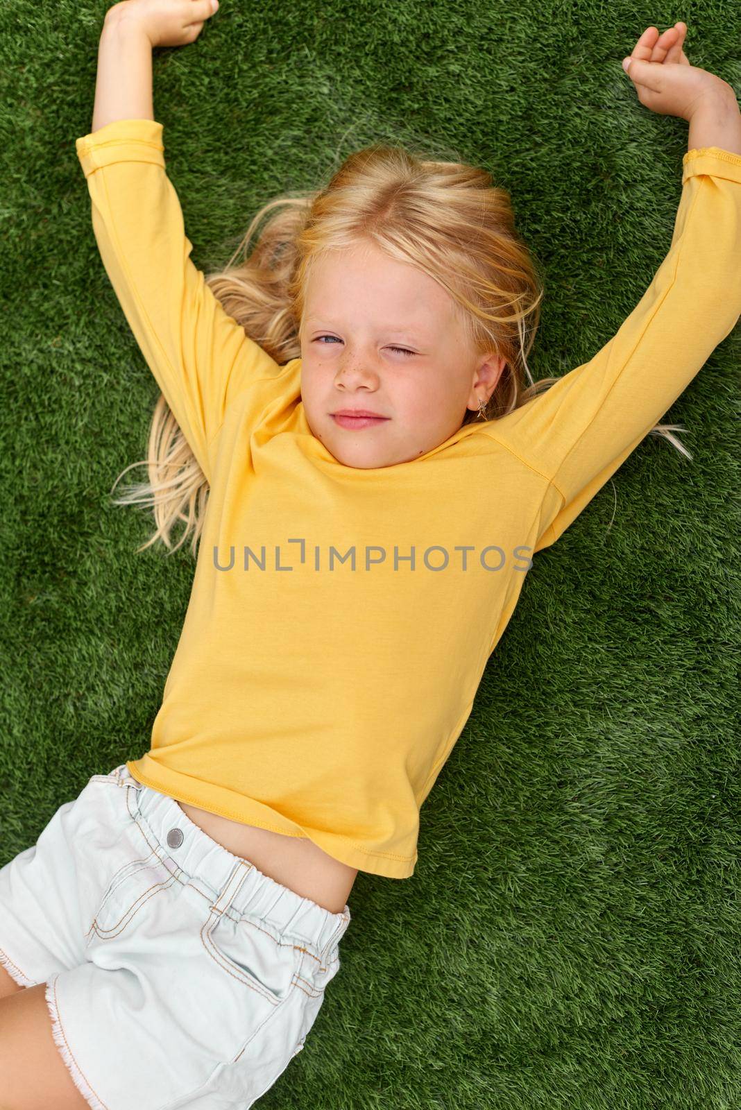Top view. Mock up for logo, text, design. Blonde long hair child girl lying, stretching on green grass. Preschool girl 5-6 years old in yellow t shirt. Lifestyle Summer vacation Leisure. People