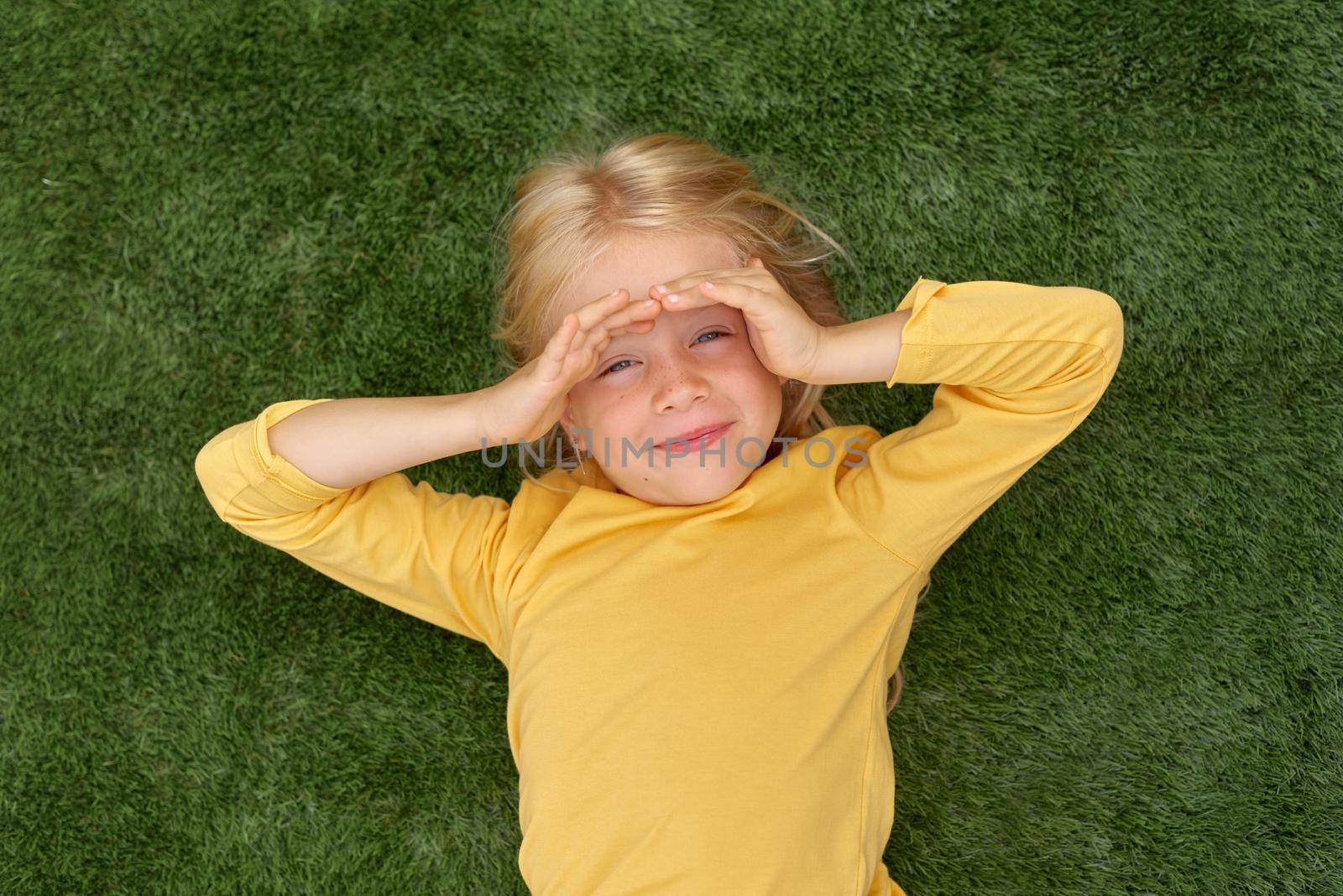 Top view. Mock up for logo, text, design. Blonde long hair child girl lying on green grass. Preschool girl 5-6 years old in yellow t shirt. Lifestyle Summer vacation Leisure. People