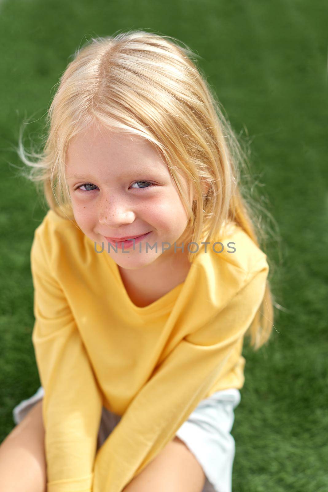Pretty beautiful blonde child girl sitting on green grass. Smiling preschool girl 5 - 6 years old in yellow t shirt looking at camera. Lifestyle. Beauty. Summer vacation. leisure. people. Top view