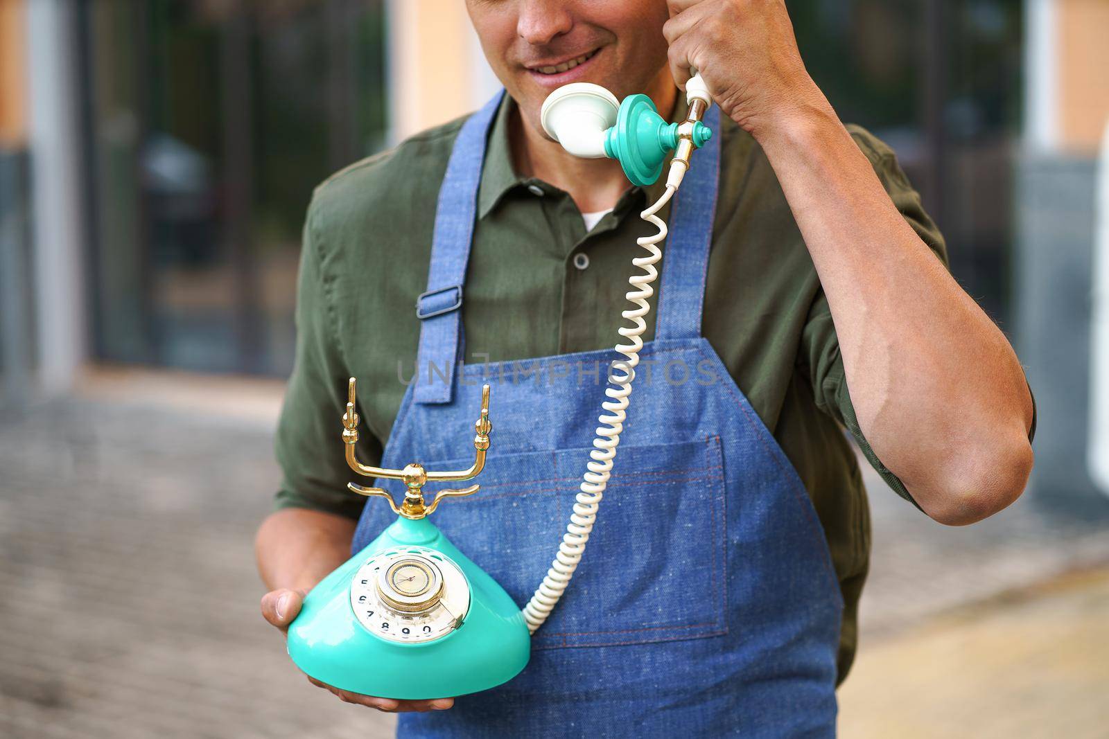 Young man talking on the vintage, old phone standing outdoors wearing apron. Service man having conversation using old phone. Telephone communications concept. No face visible.