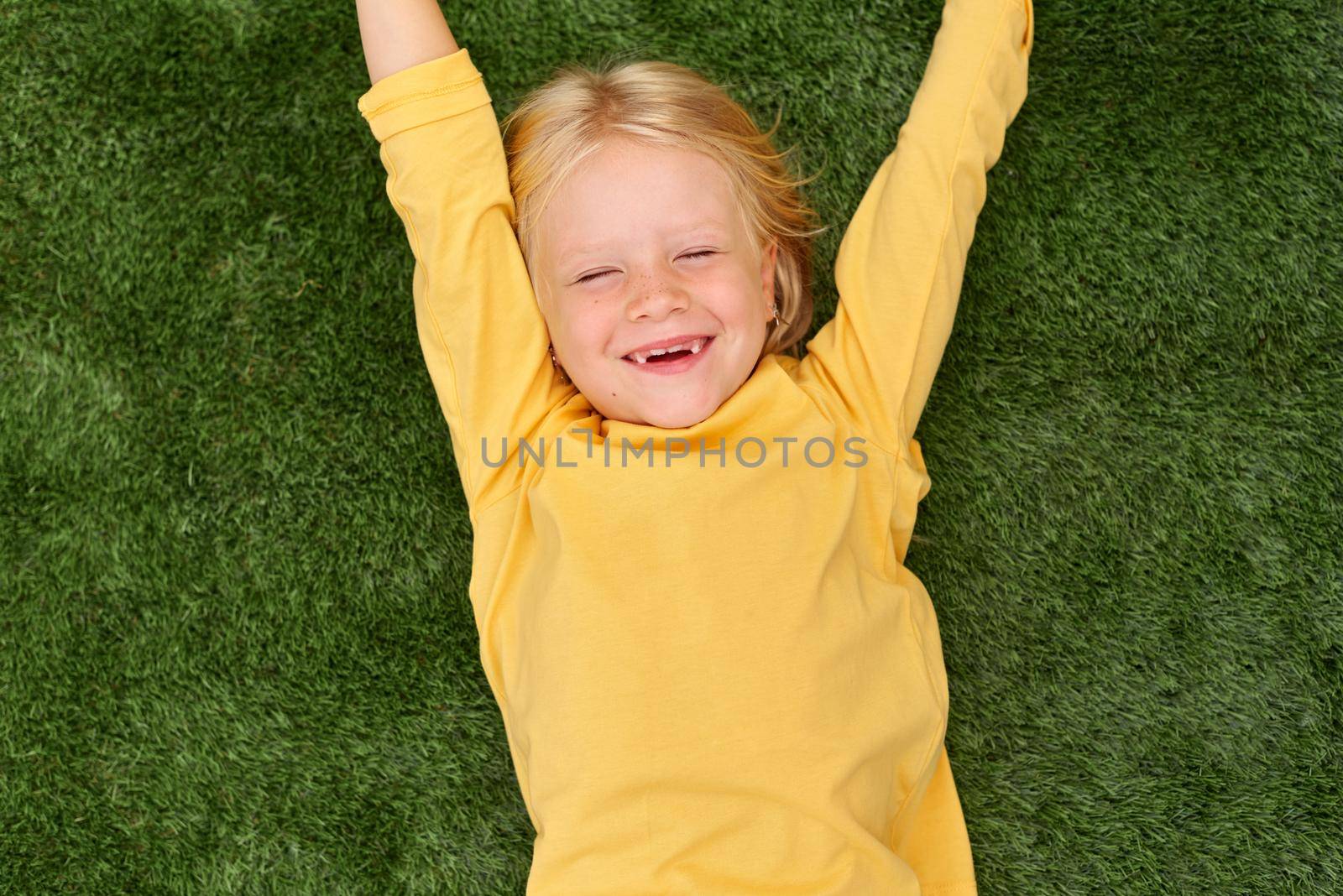 Top view. Mock up for logo, text, design. Blonde long hair child girl lying, stretching on green grass. Preschool girl 5-6 years old in yellow t shirt. Lifestyle Summer vacation Leisure. People