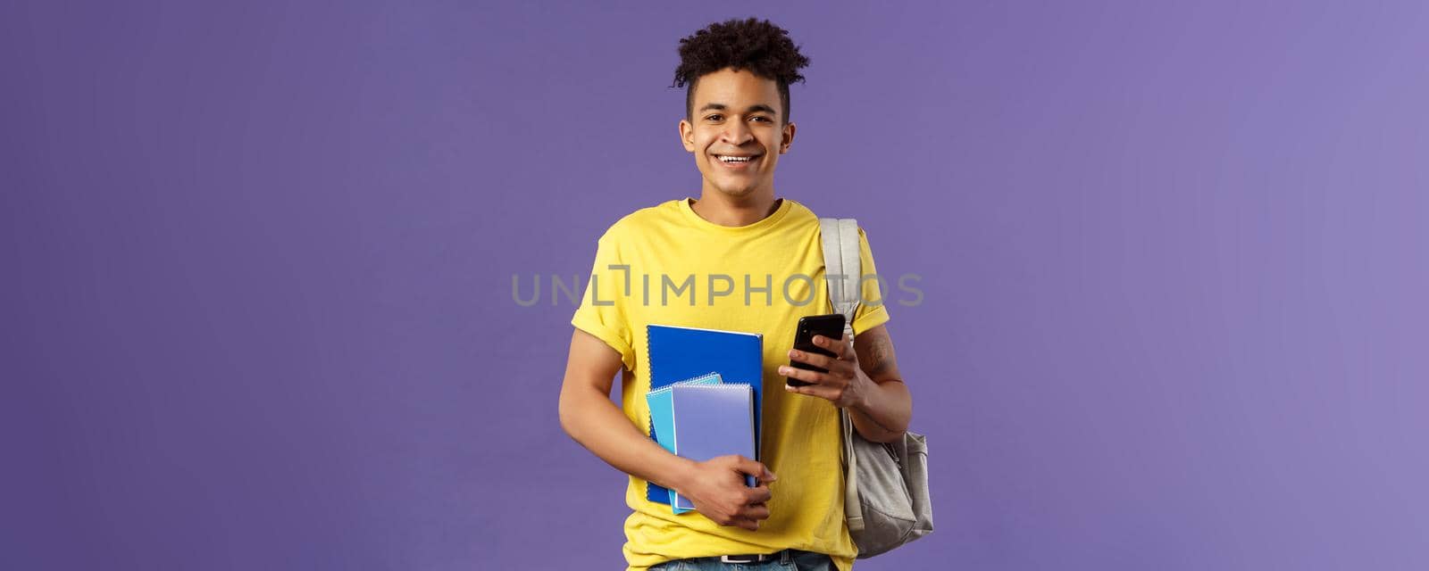 Back to school, university concept. Portrait of cheerful young handsome male student wearing backpack on shoulder, hold notebooks and studying books, texting friend mobile phone.