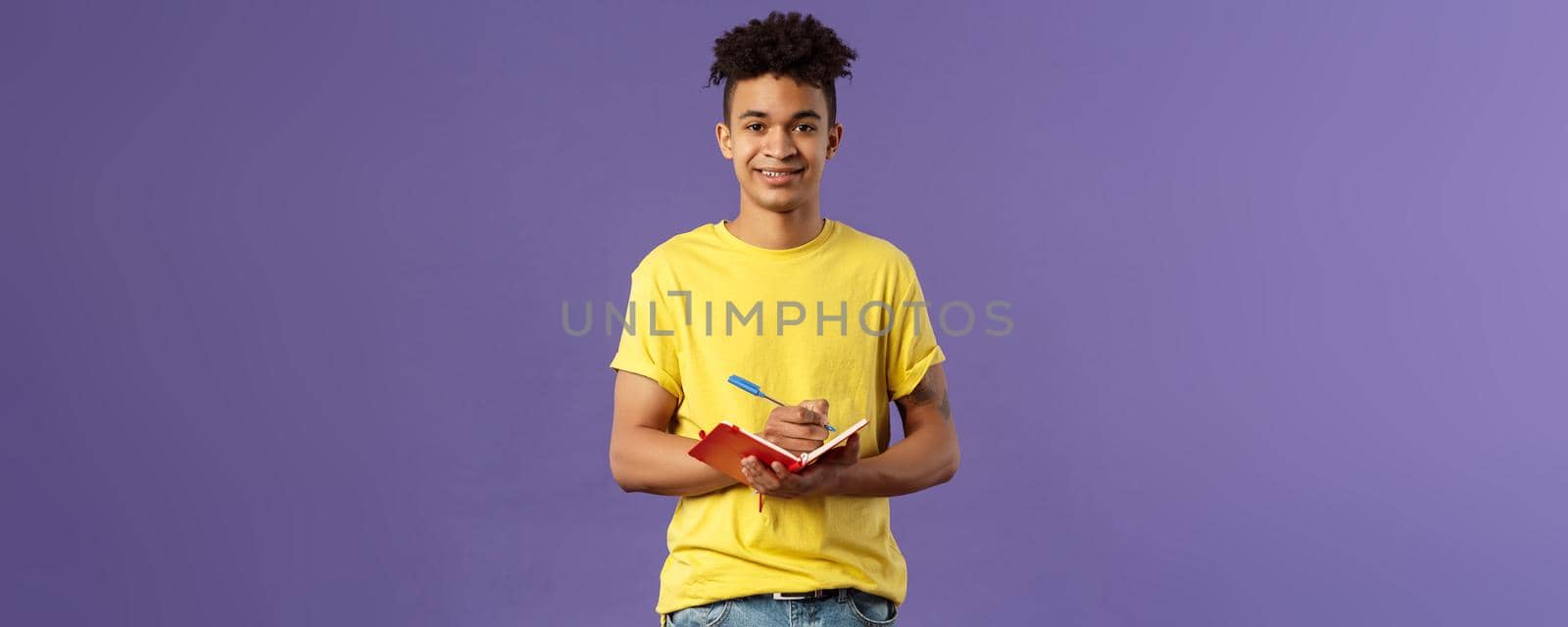 Portrait of young hispanic male student studying online courses, writing down lecture, making personal schedule or taking notes in planner, look camera enthusiastic and interested, purple background