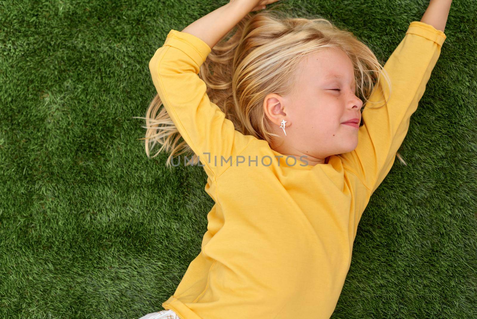 Portrait cute blonde girl with closed eyes lying on green grass. Mock up yellow t shirt. Top view. by OlgaPink