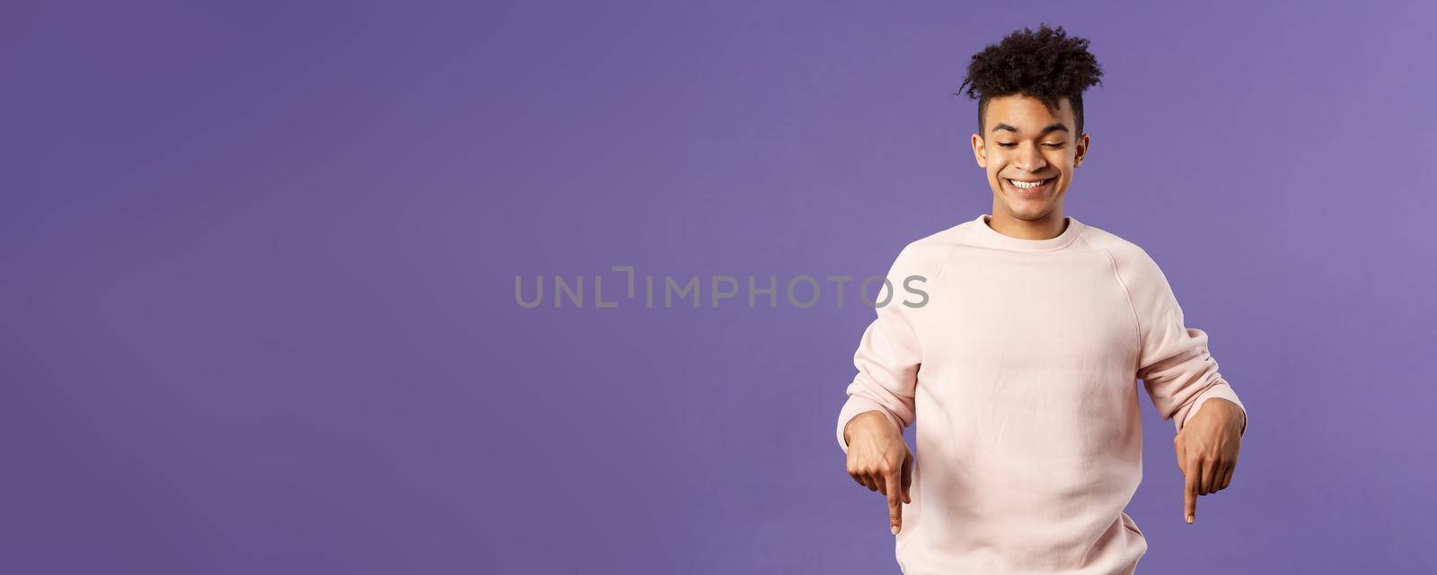 Portrait of cute enthusiastic young man found something really cool, pointing fingers down, happy smiling and peeking at bottom advertisement with pleased grin, purple background.