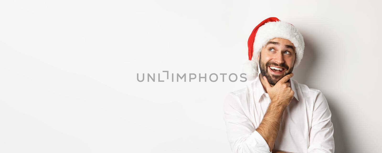 Party, winter holidays and celebration concept. Close-up of happy man planning christmas gift list, wearing santa hat, looking at upper left corner thoughtful, white background.