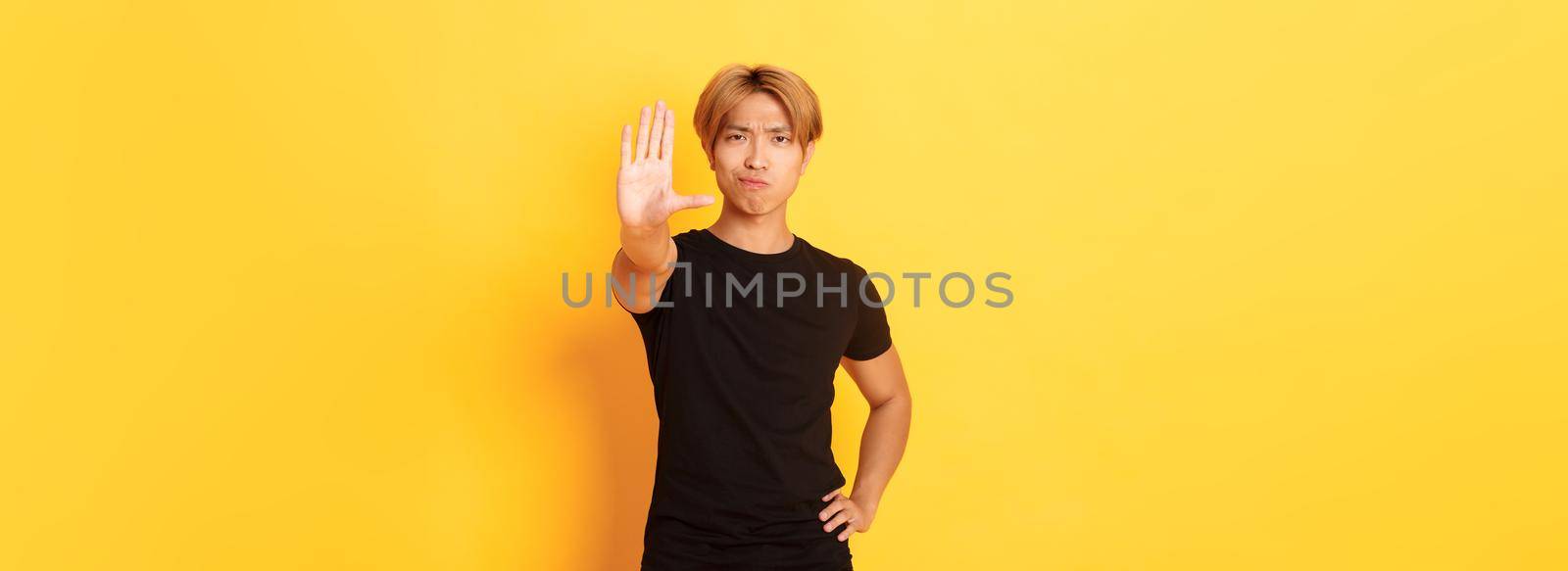 Portrait of disappointed serious asian man, smirking displeased and extend hand, showing stop gesture, yellow background by Benzoix