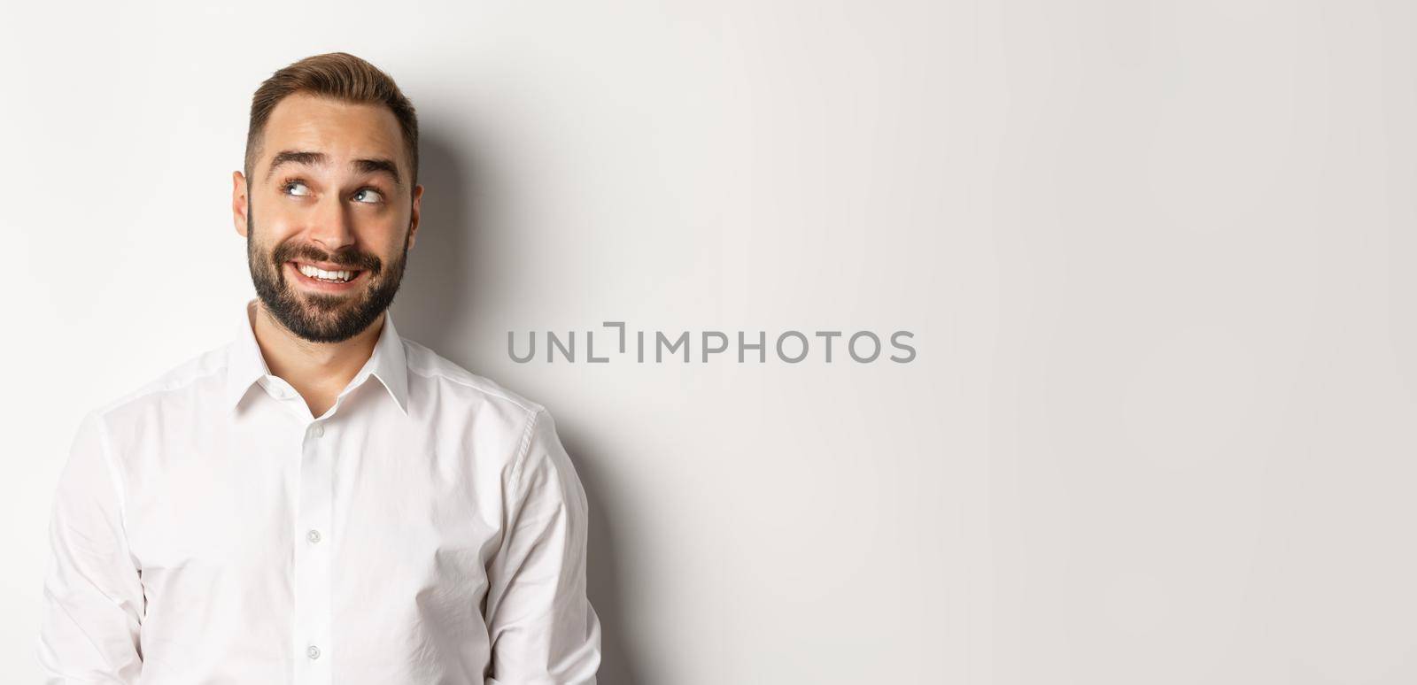 Close-up of handsome man with beard, looking thoughtful at upper left corner, imaging and smiling, white background.