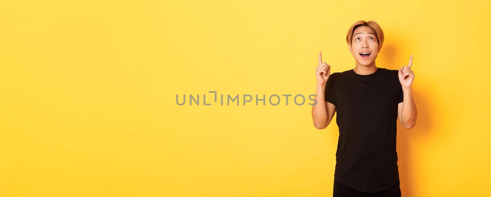 Impressed good-looking asian blond guy, wearing black t-shirt, looking and pointing fingers up excited, yellow background.