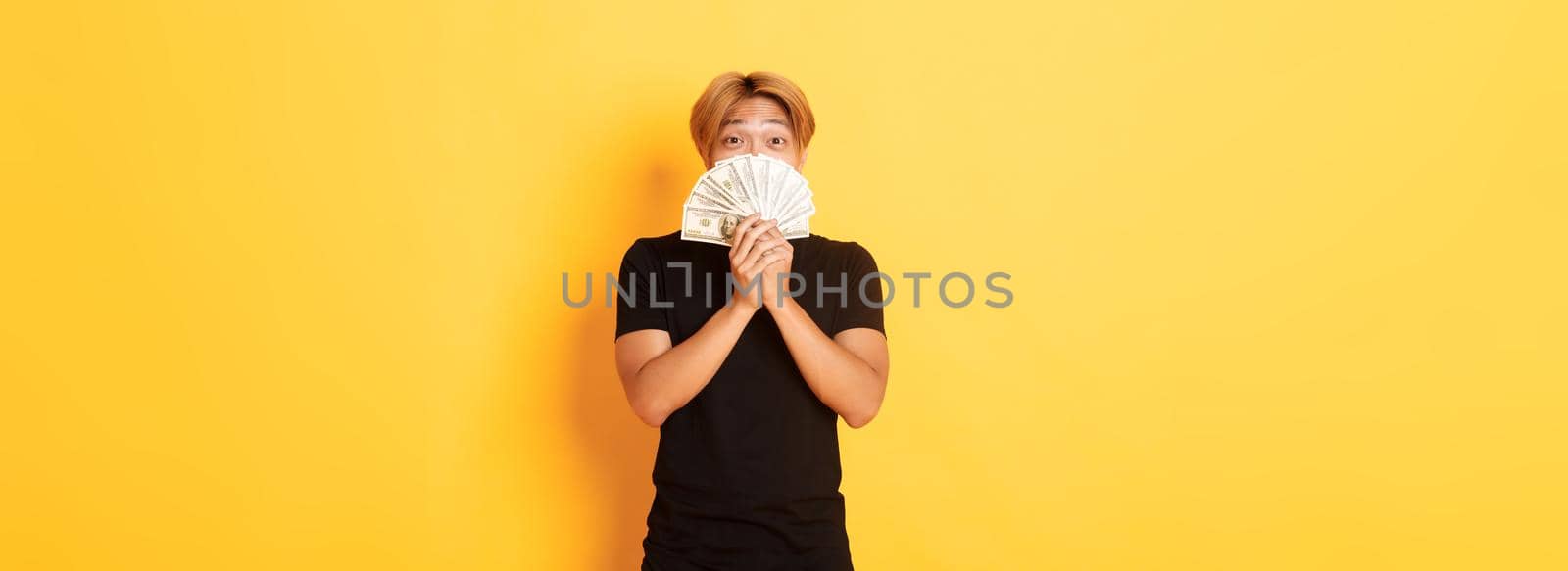 Excited lucky blond asian guy rejoicing from winning cash, holding money and looking happy, standing yellow background by Benzoix