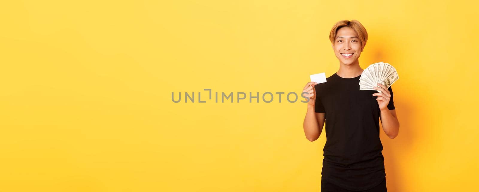 Pleased handsome asian guy showing money and credit card, smiling happy, standing yellow background by Benzoix