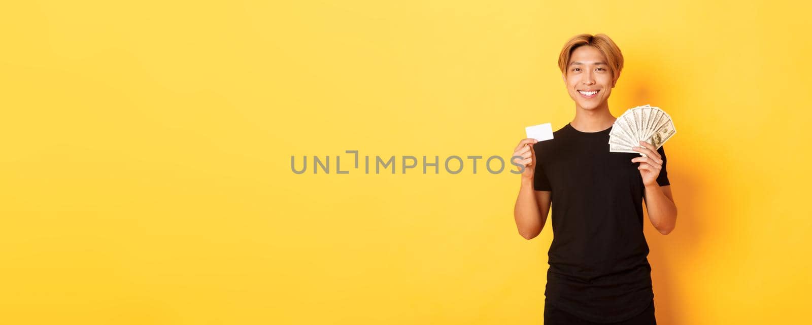 Pleased handsome asian guy showing money and credit card, smiling happy, standing yellow background.