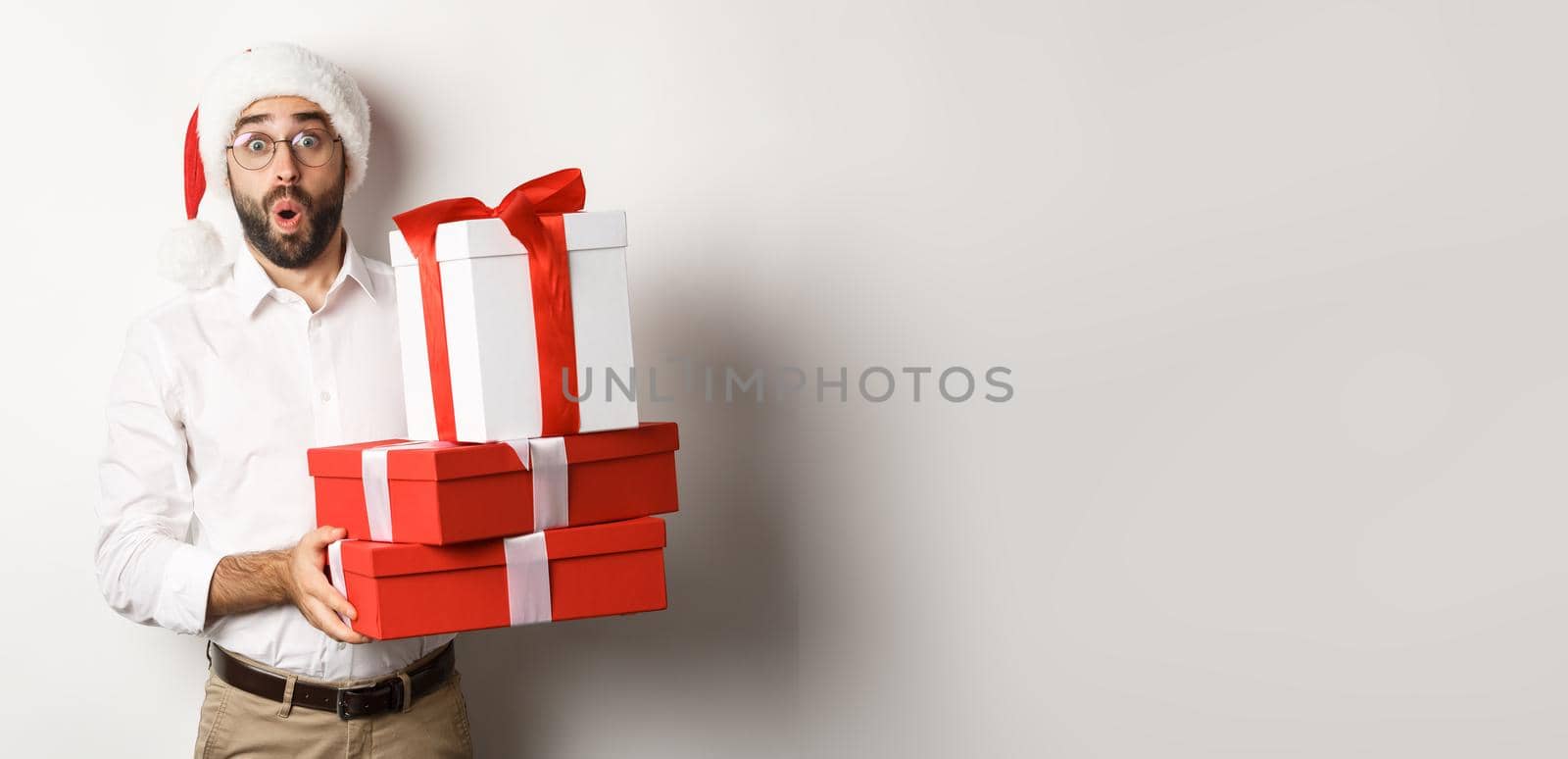 Winter holidays and celebration. Excited man holding Christmas gifts and looking surprised, wearing Santa hat, standing over white background.
