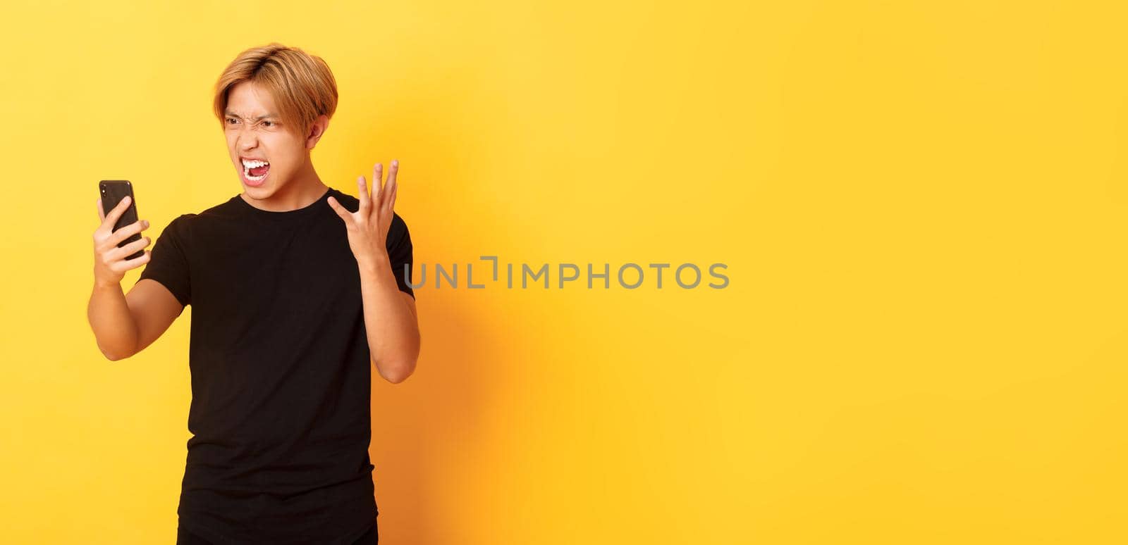 Portrait of angry and pissed-off asian man looking mad at smartphone screen, having argument during video call, standing yellow background by Benzoix