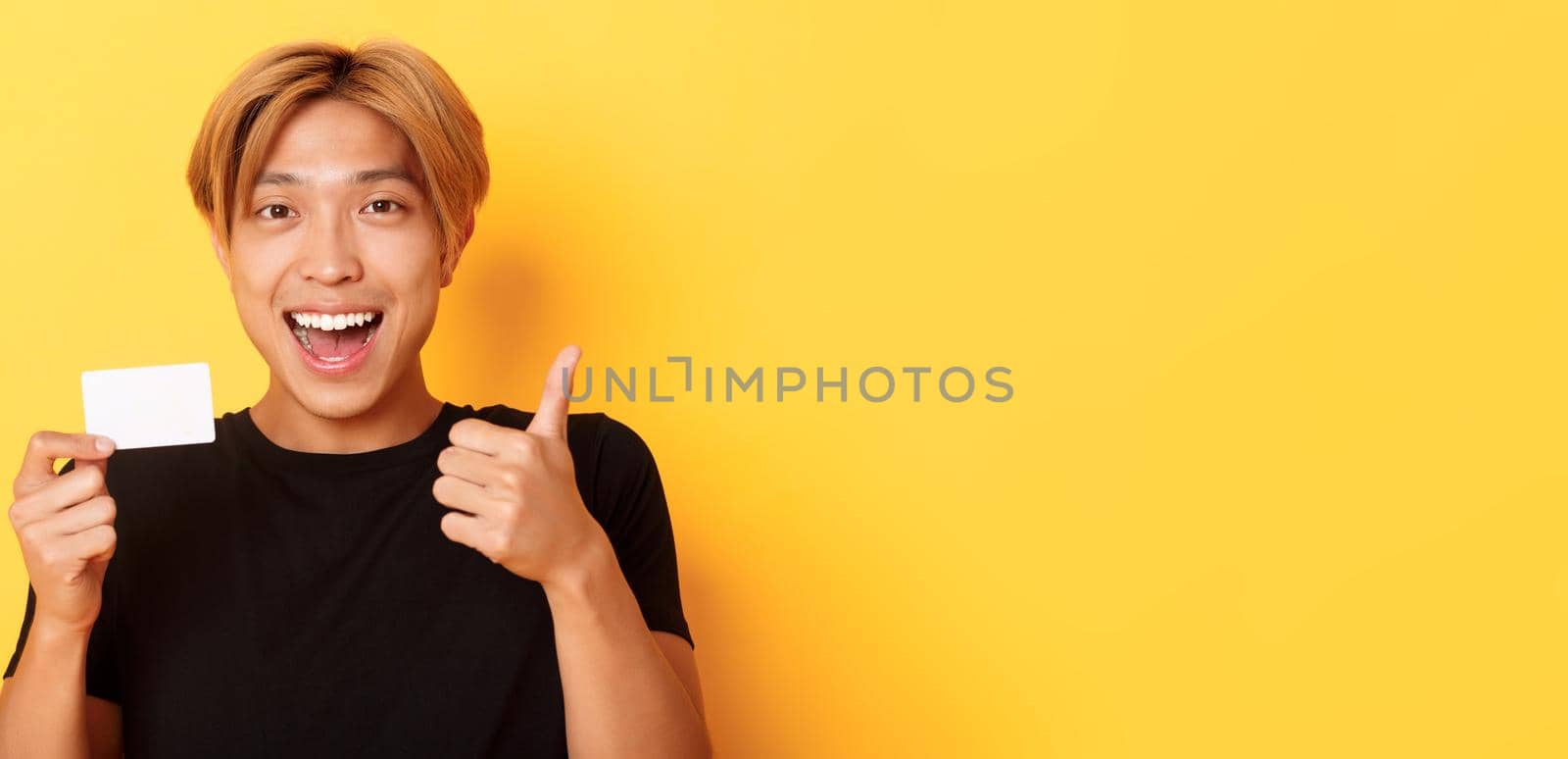 Close-up of happy and satisfied asian handsome guy, showing credit card and thumbs-up in approval, smiling amazed, standing yellow background by Benzoix