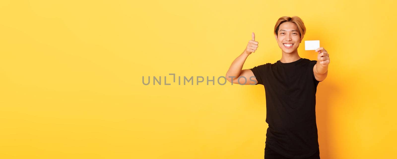 Satisfied handsome asian guy smiling pleased and showing credit card, thumbs-up in approval, standing yellow background by Benzoix