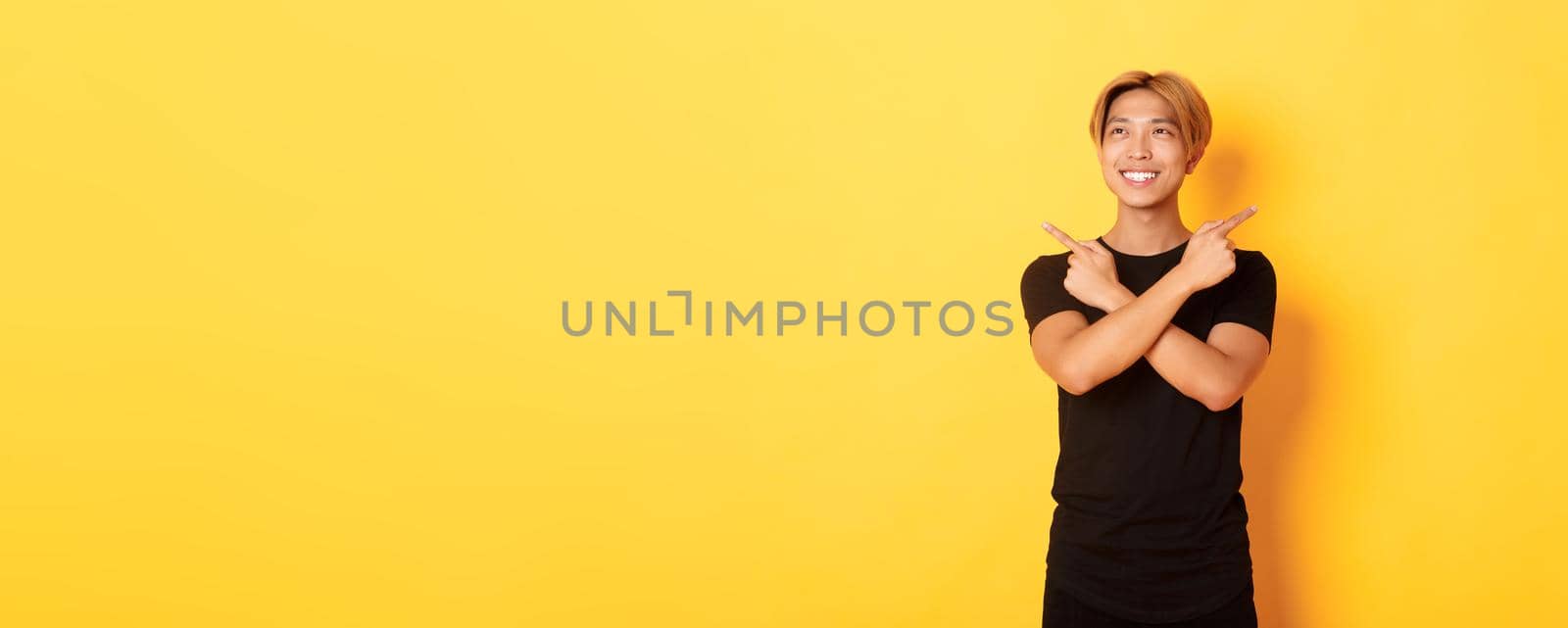 Portrait of smiling pleased asian guy, looking left and making decision, pointing sideways, yellow background by Benzoix