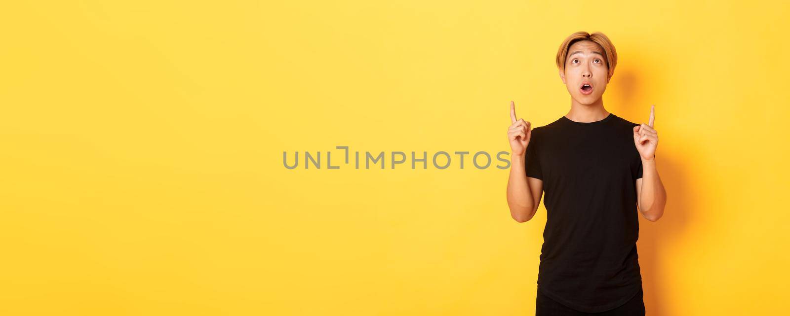 Portrait of curious and amazed asian guy with blond hair, wearing black t-shirt, looking and pointing fingers up astonished, yellow background.