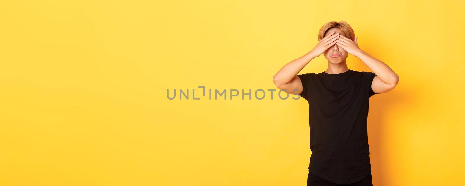 Portrait of sad and miserable asian guy shut eyes and sulking upset, standing yellow background.