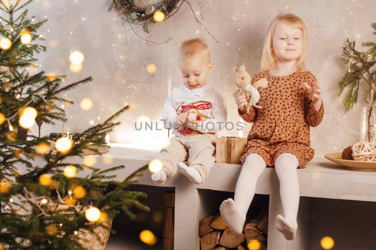 Little brother and sister play on Christmas eve in a beautiful house decorated for the New Year holidays. Children are playing with a Christmas gift. Scandinavian-style interior with live fir trees and a wooden staircase.