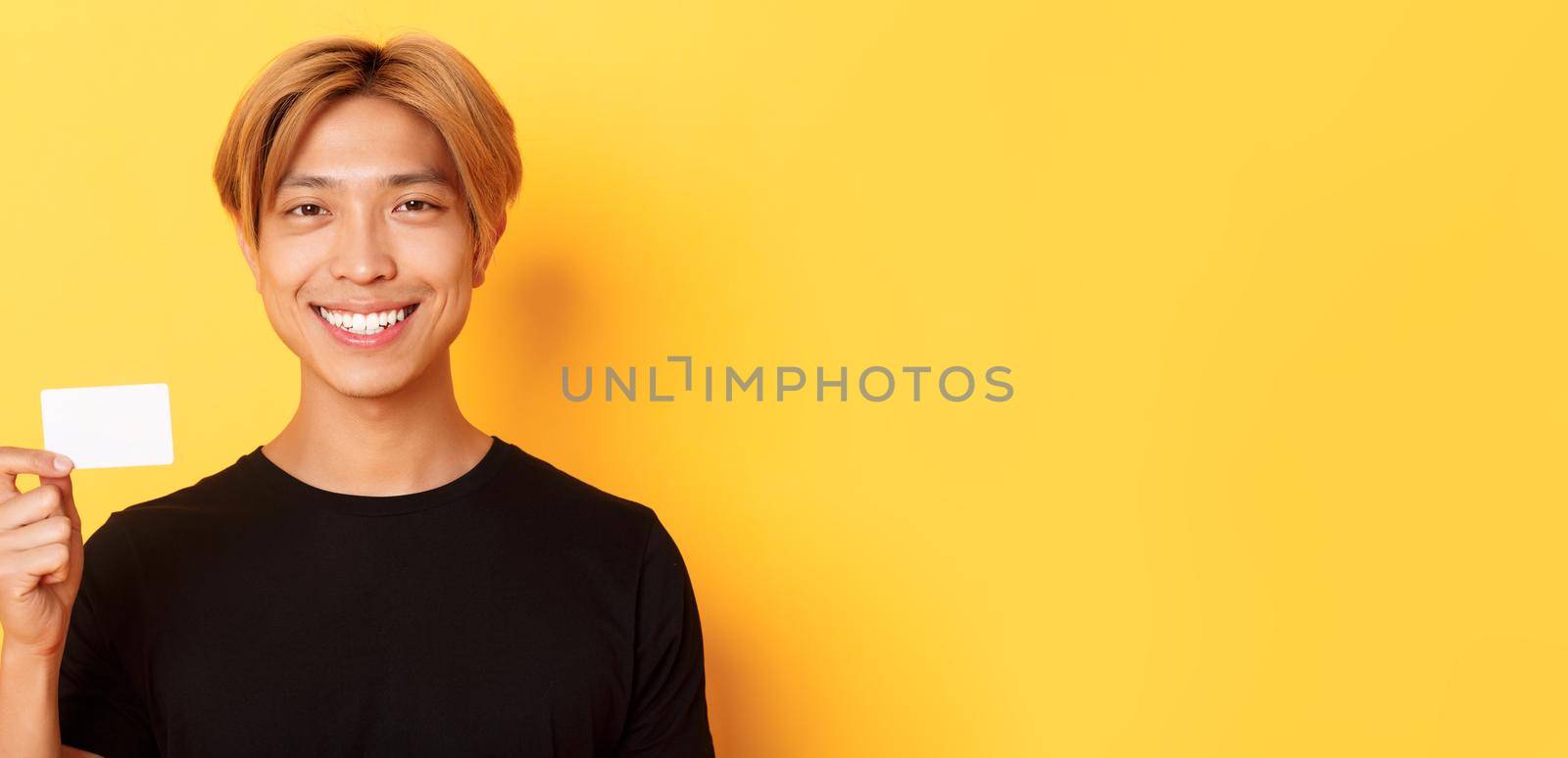Close-up of handsome pleased asian guy with fair hair, smiling happy and showing credit card over yellow background by Benzoix