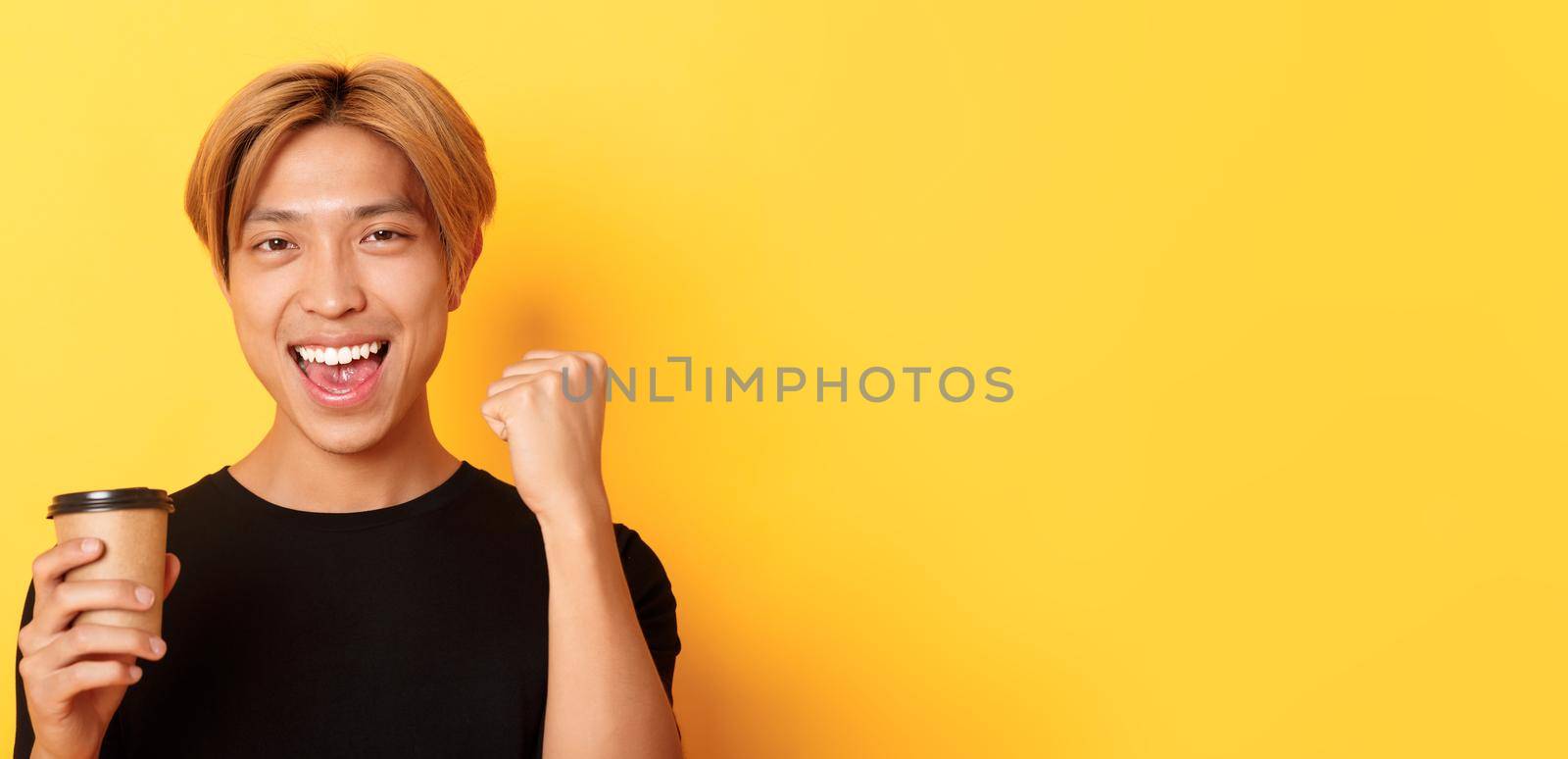 Close-up of energized handsome asian guy fist pump joyfully while drinking coffee, smiling excited over yellow background by Benzoix