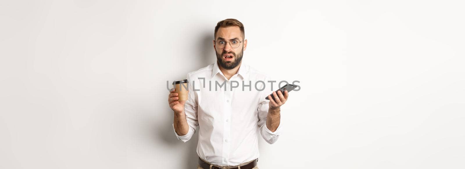 Image of man drinking coffee, feeling confused about strange message on mobile phone, standing over white background.