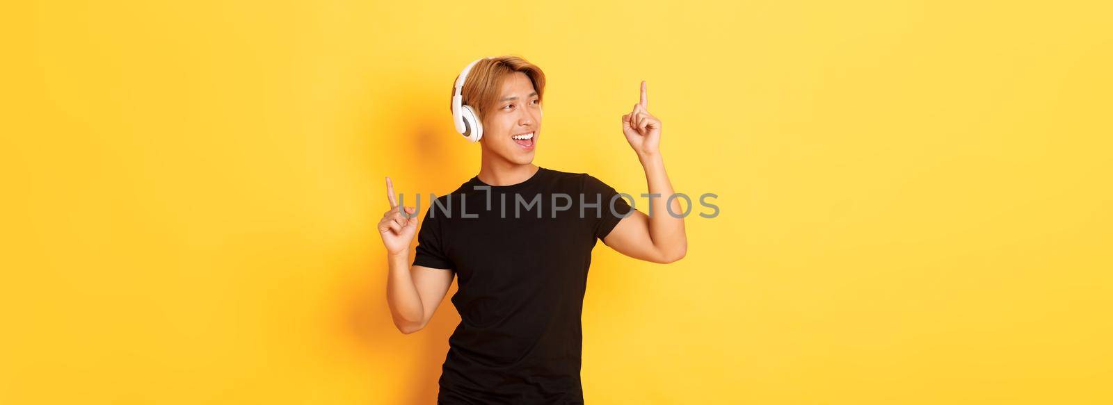 Joyful handsome asian guy with blond hair, singing along and dancing as listening to music in wireless headphones, standing yellow background by Benzoix
