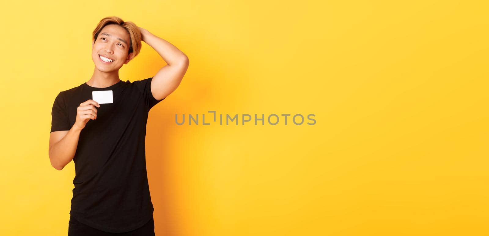Thoughtful smiling asian guy thinking while showing credit card, looking upper left corner dreamy, yellow background by Benzoix