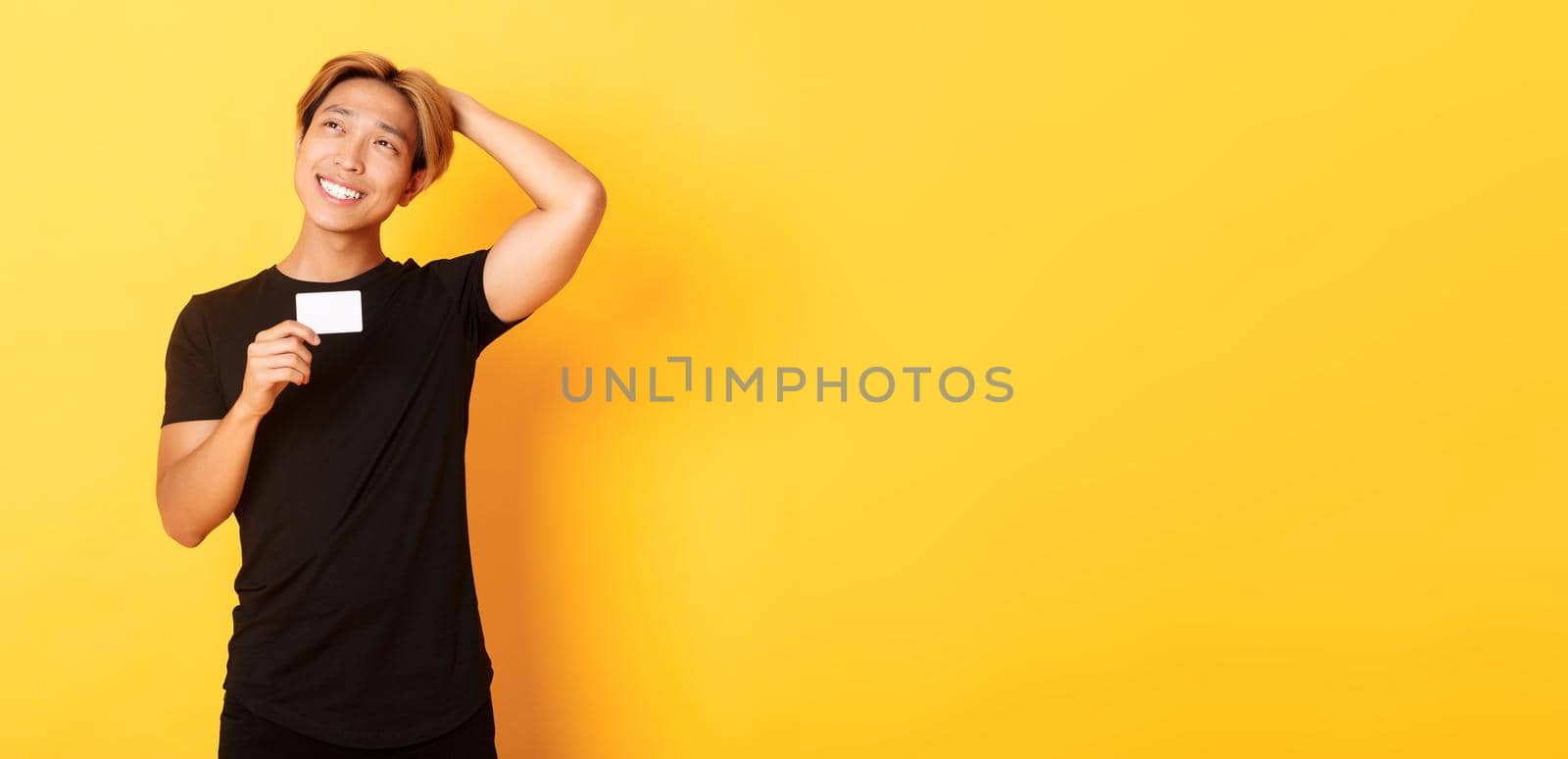 Thoughtful smiling asian guy thinking while showing credit card, looking upper left corner dreamy, yellow background.