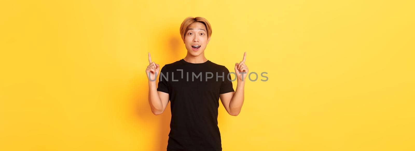 Portrait of surprised and excited asian guy looking amused, pointing fingers up, yellow background.