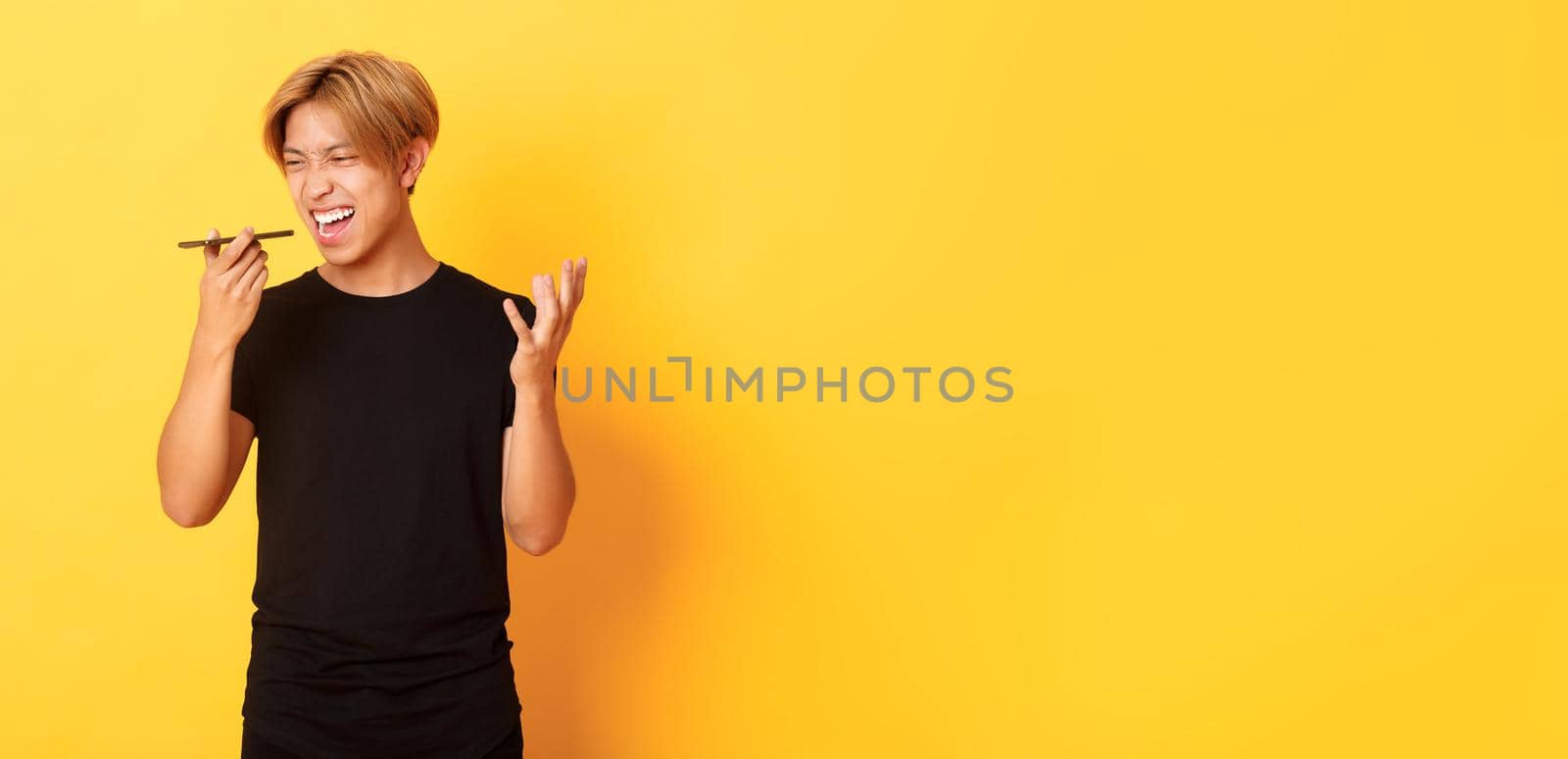 Portrait of angry asian man yelling mad at mobile phone speaker, record furious voice message, standing yellow background.