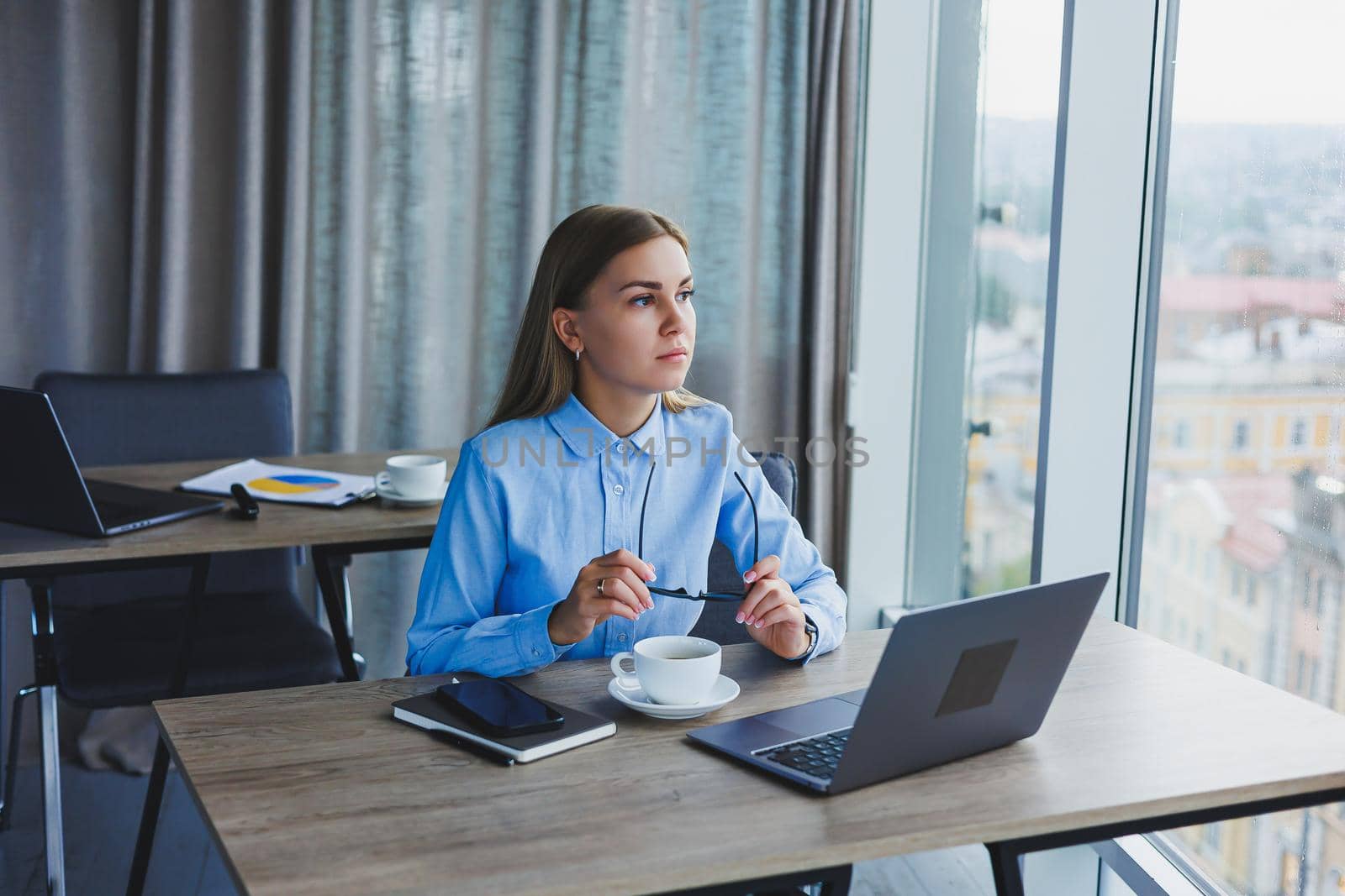 Adult smiling female manager in glasses and formal shirt sits at a desk and looks at a laptop. Modern, spacious, bright office with a large window.