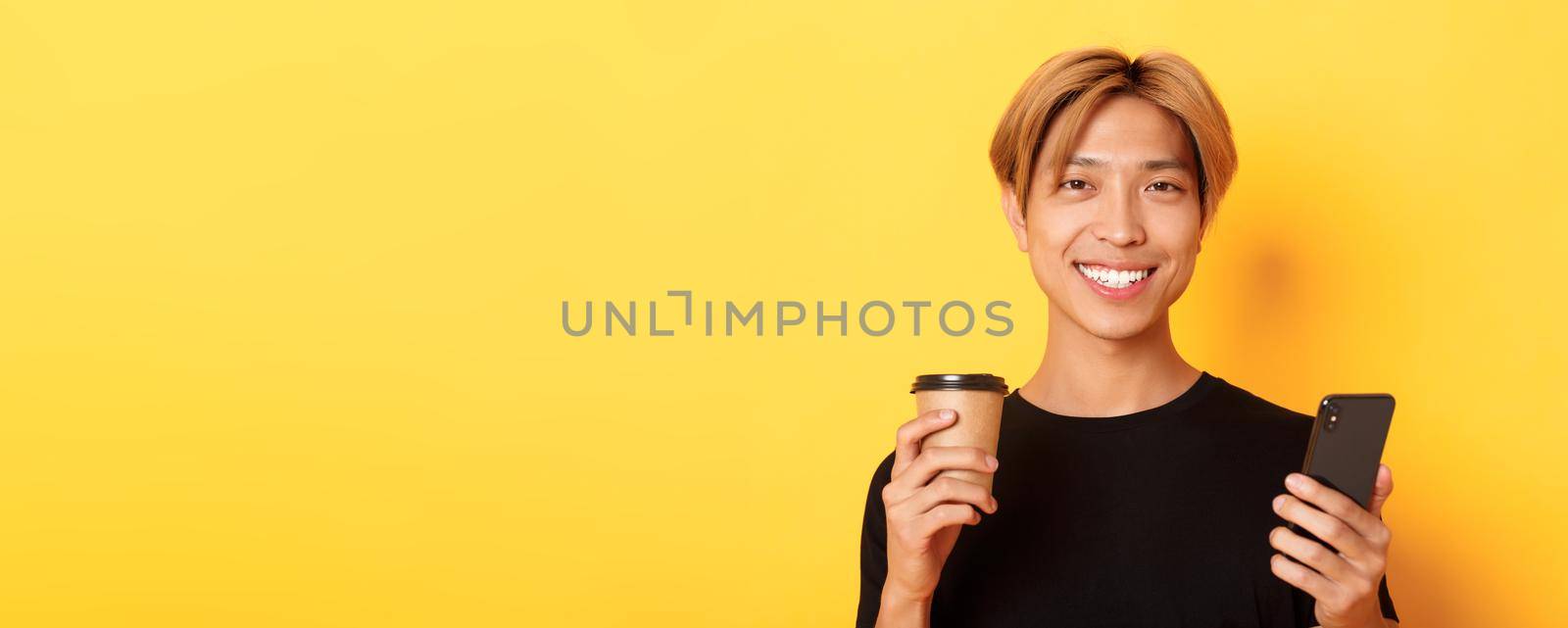 Close-up of handsome young asian guy smiling happy, using smartphone and drinking takeaway coffee, standing over yellow background by Benzoix