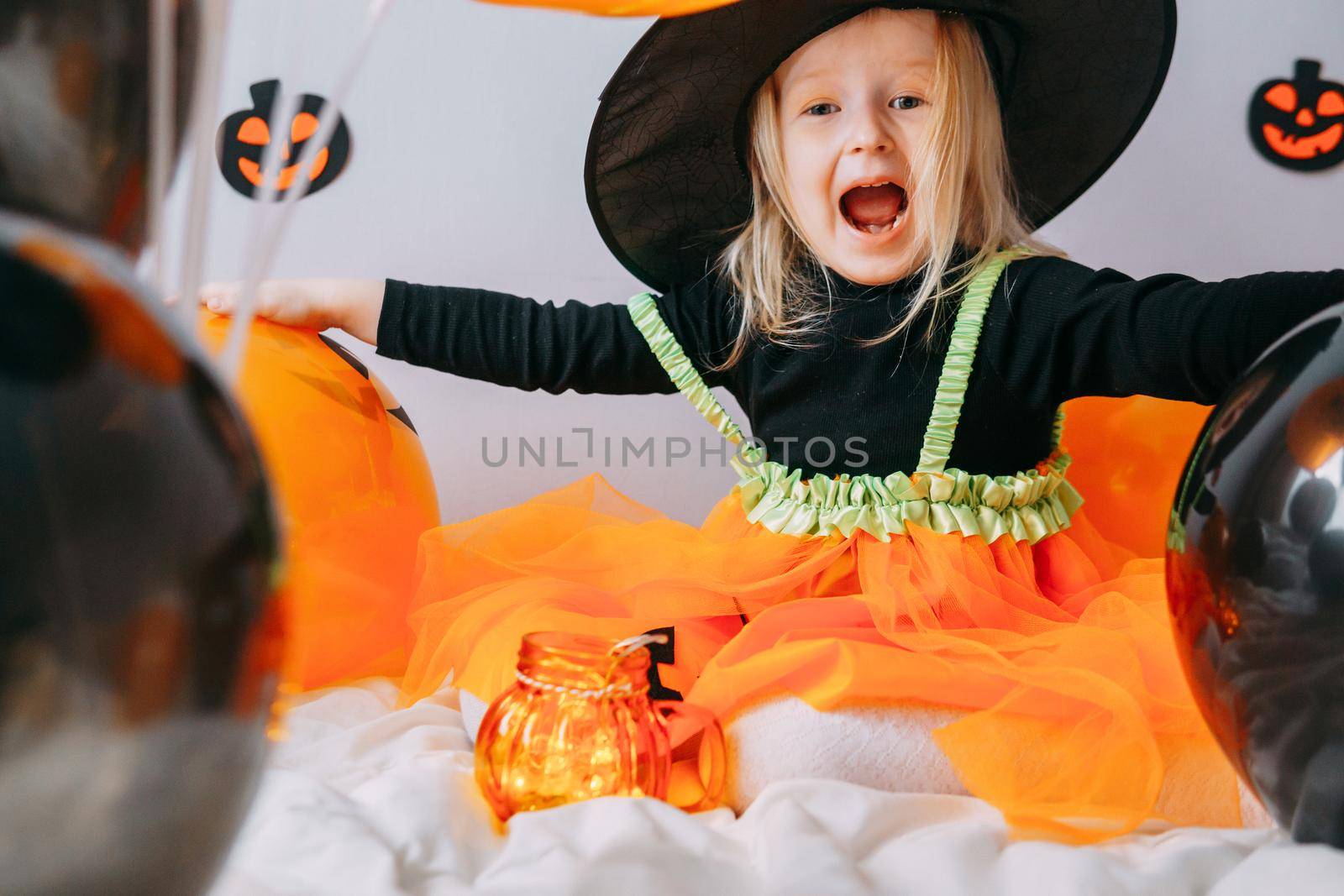 Children's Halloween - a girl in a witch hat and a carnival costume with airy orange and black balloons at home. Ready to celebrate Halloween.