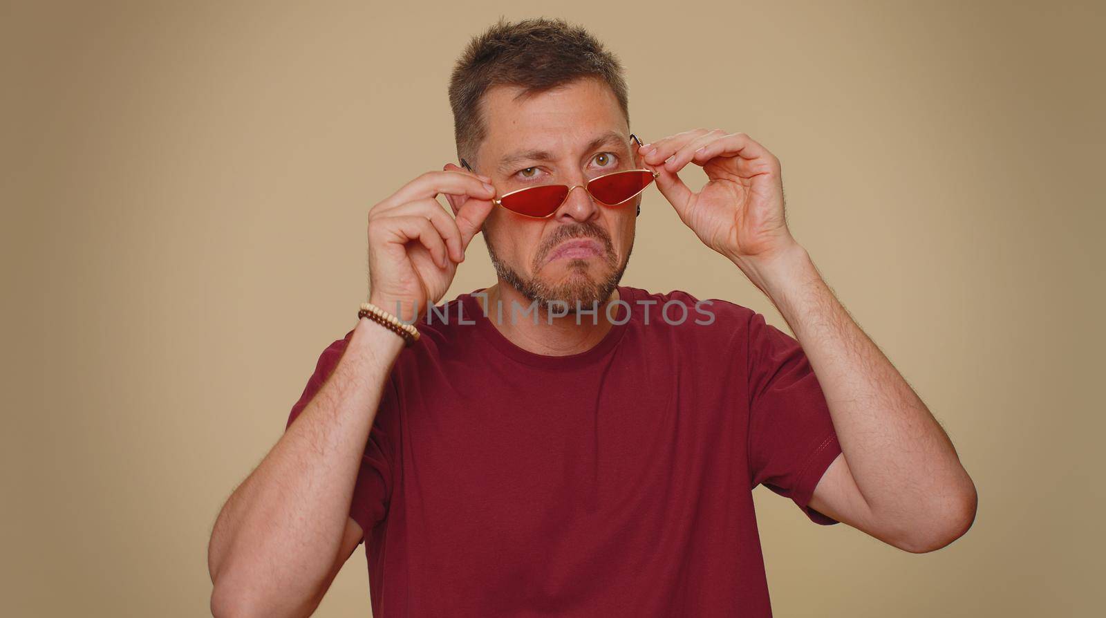 Cheerful bearded handsome young man in t-shirt and red sunglasses smiling, looking at camera. Adult stylish positive male hipster guy fashion model isolated alone on beige studio background indoors