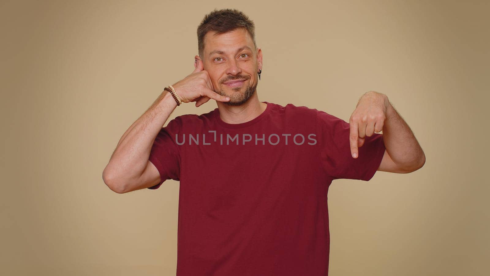 Call me, here is contact number. Bearded handsome young man looking at camera doing phone gesture like says hey you call me back. Adult stylish male guy isolated on beige studio background indoors