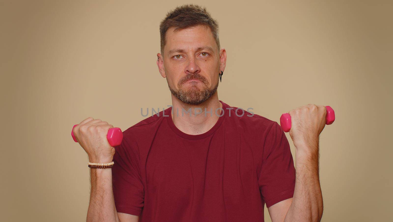 Handsome young man in red t-shirt working out, pumping up arm muscles lifting pink gym dumbbells, practicing pilates, healthcare. Adult stylish guy on beige studio wall background. Sport motivation