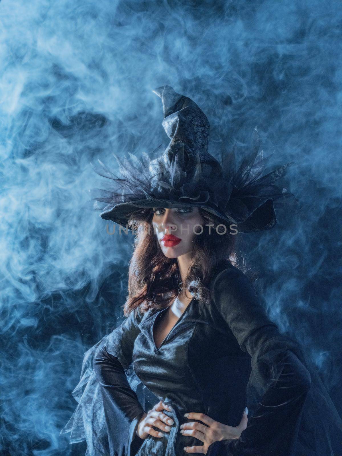 Beautiful woman in Halloween witch costume dress and hat in mist, blue mysterious light