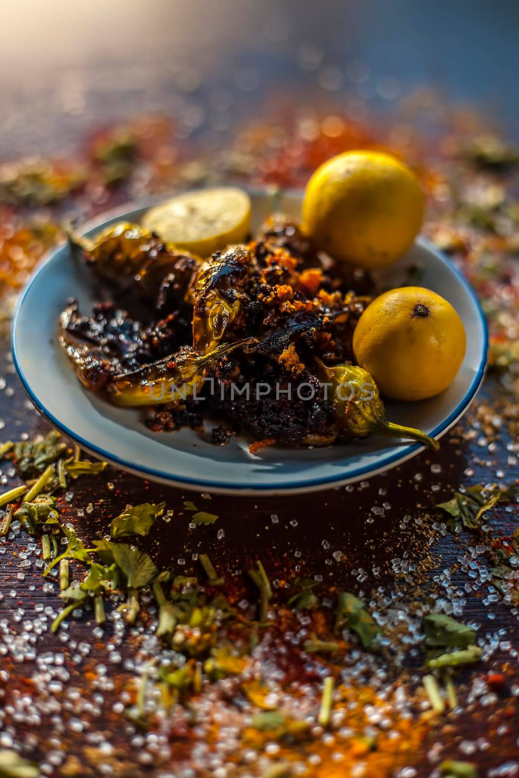Close up shot of Classic Bharela Marcha or Stuffed fried chilies in a glass plate along with lemons and all the spices needed for preparing it on a brown wooden surface. by mirzamlk