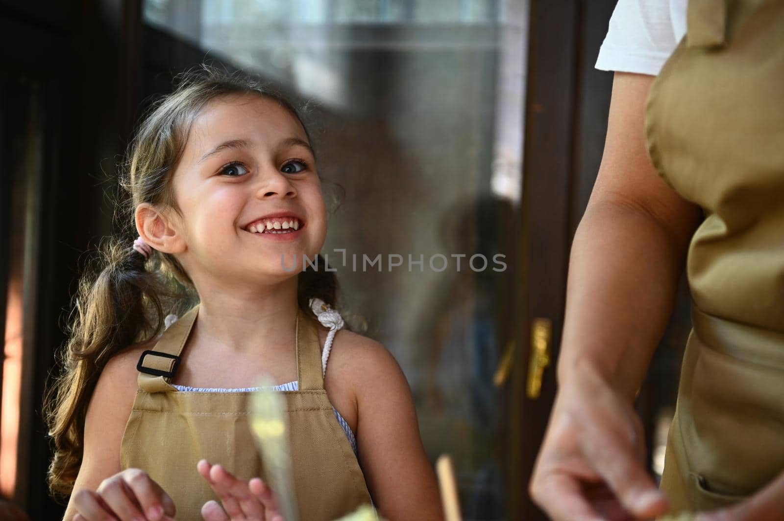 Adorable beautiful Caucasian child with two ponytails little girl in a beige chef's apron, smiling a cheerful toothy smile, feeling happiness while cooking dinner with her mom in the country kitchen