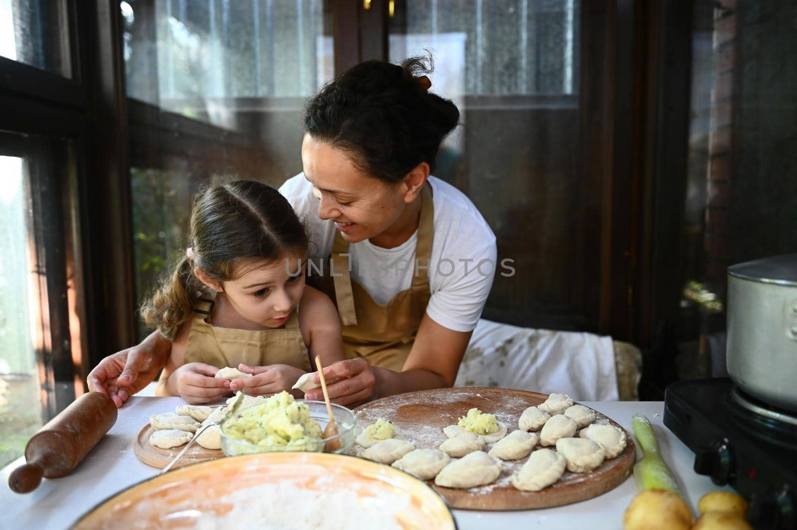 Loving mother hugging her adorable little daughter, standing together at a table with dumplings on a wooden board, made according to traditional Ukrainian recipe, enjoying together cooking varenyky