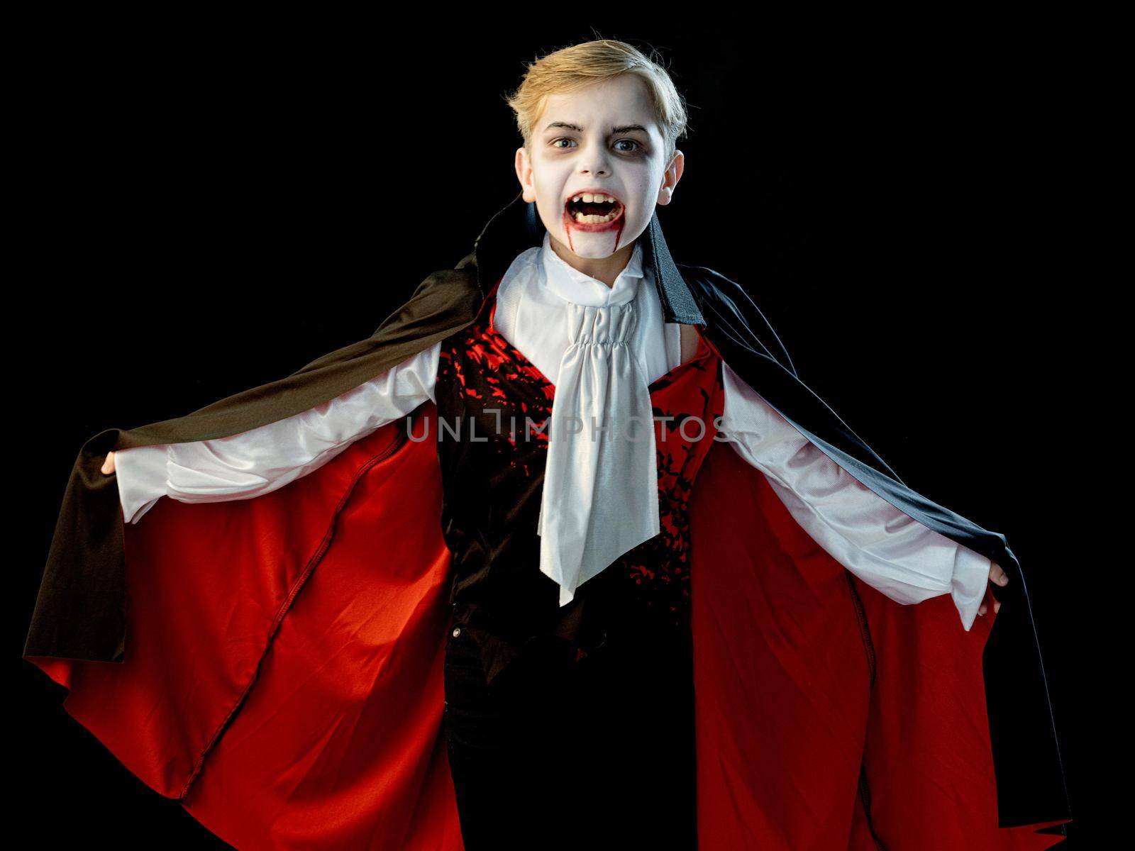 Portrait of boy wearing Halloween vampire makeup and costume cloak bared his teeth, isolated on black background