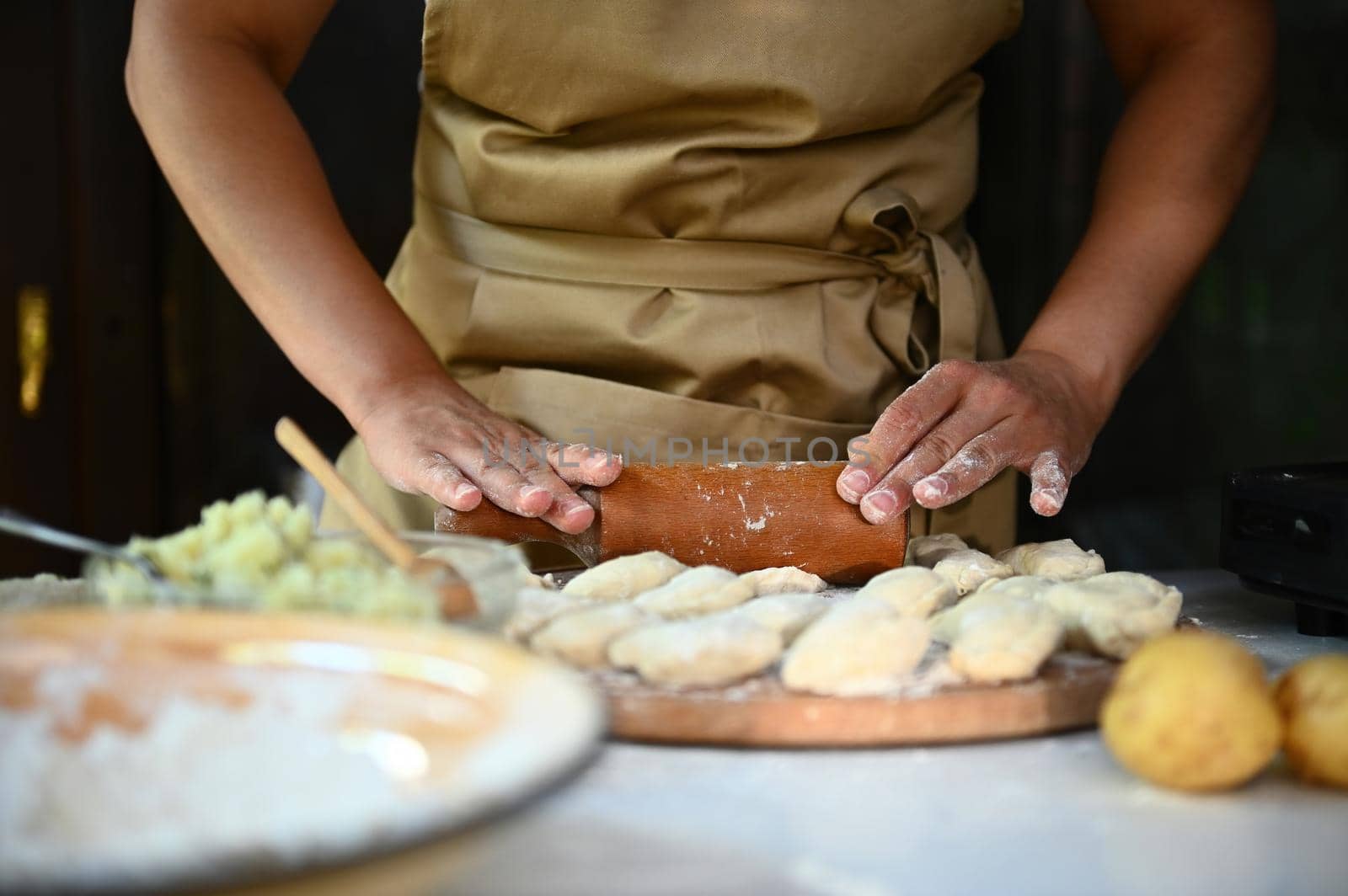 Close-up woman chef housewife in beige apron, rolls out the dough for dumpling with rolling pin, makes homemade vareniki by artgf